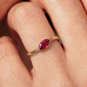 Op de afbeelding: Een gouden ring met een enkele dieprode edelsteen in marquise-slijpvorm, horizontaal in het midden gezet. De ringband is glad en gepolijst, met een eenvoudig ontwerp dat de gefacetteerde robijnkleurige steen in kleine metalen klauwen benadrukt.