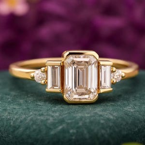 Könnte beinhalten: Ein goldener Ring mit einem smaragdgeschliffenen Mittelstein, flankiert von zwei Baguette-Steinen und kleinen runden Akzentsteinen. Der Ring ist auf einem goldenen Band gefasst. Verschwommener violetter Hintergrund.