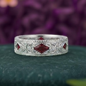 May include: A silver band ring featuring a repeating pattern of engraved floral motifs and alternating kite-shaped red gemstones. The metal surface displays intricate detailing with a textured border along the top and bottom edges of the ring.