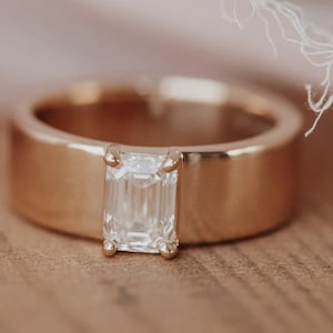 May include: A gold band ring featuring a rectangular, clear gemstone. The ring is set on a wooden surface, with a soft pink fabric and white decorative element in the background. The ring's design is simple and elegant.