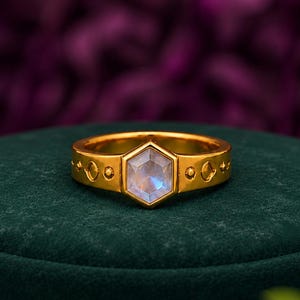 May include: A gold-toned ring featuring a central hexagonal-cut moonstone with a soft blue iridescent sheen. The band is crafted with a polished finish and includes engraved geometric patterns consisting of small circles and diamond shapes.