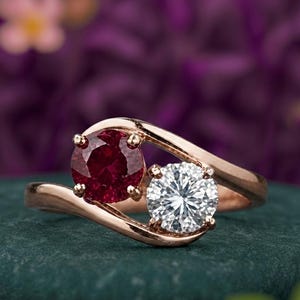 Puede incluir: Un anillo tipo bypass de metal en tono rosado con dos piedras de corte redondo. Una piedra es de color rojo intenso, mientras que la otra es una piedra transparente, incolora y facetada. La banda se curva alrededor de ambas piedras.