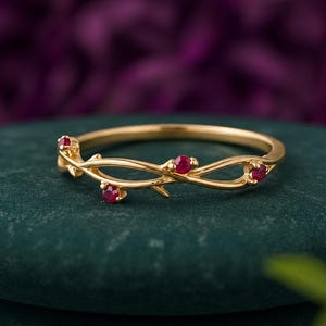 Puede incluir: Un anillo de oro con un diseño delicado de banda entrelazada similar a una enredadera. La banda está acentuada con tres pequeñas piedras preciosas redondas de color rojo vibrante, engastadas a intervalos a lo largo de la estructura metálica.