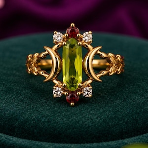 May include: A gold ring featuring a central vertical baguette-cut lime green gemstone flanked by two crescent moon shapes. The setting includes four small clear round stones and two small dark red round stones at the top and bottom points.