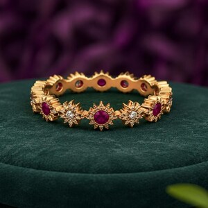 May include: A gold band ring featuring an alternating pattern of round red gemstones and star-shaped settings containing small clear stones. The ring has a continuous design of sunburst-like motifs that encircle the entire circumference of the band.