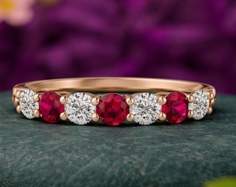 May include: A gold band ring featuring alternating round-cut rubies and diamonds. The ring has a classic design with the gemstones set closely together. The ring is displayed on a dark green stone against a blurred purple background.
