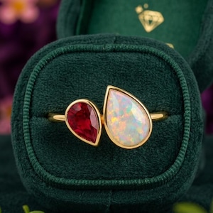 May include: A gold ring featuring two pear-shaped gemstones set side-by-side. One stone is a deep red ruby, and the other is a white opal with iridescent flashes of color. The ring is displayed within a dark green velvet jewelry box.