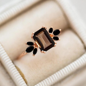 May include: A rose gold ring featuring a rectangular, smoky brown gemstone as the centerpiece. The ring is accented with black, marquise-cut stones on either side, creating a floral-like design. The ring is set in a white velvet box.