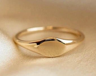 May include: A gold signet ring with a smooth, polished surface. The ring features a rounded band and a flat, oval-shaped face. The ring is set against a soft, neutral background.