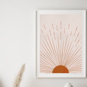 May include: A framed art print featuring a minimalist sunburst design in shades of orange and peach. The artwork hangs above a wooden cabinet with an oval shape and two small ceramic vases. A glass jar with dried pampas grass completes the decor.