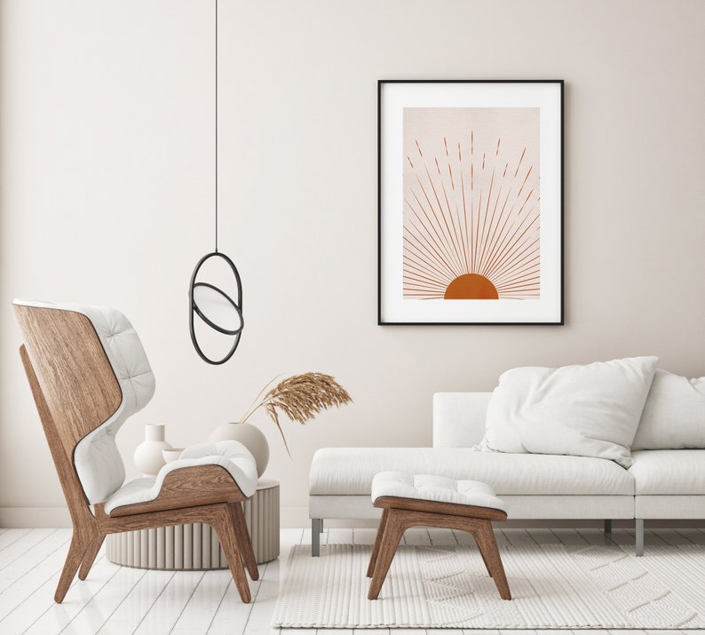 May include: A framed art print featuring a minimalist sunburst design in shades of orange and peach. The artwork is displayed above a white sofa and wooden chair, creating a warm and inviting living room setting. A modern black pendant light hangs from the ceiling.
