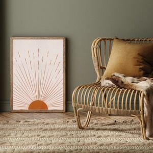May include: A framed art print featuring a minimalist sunburst design in shades of orange and beige. The artwork is displayed next to a woven rattan chair with a mustard-colored pillow and a faux fur throw. A beige rug and green wall complete the scene.