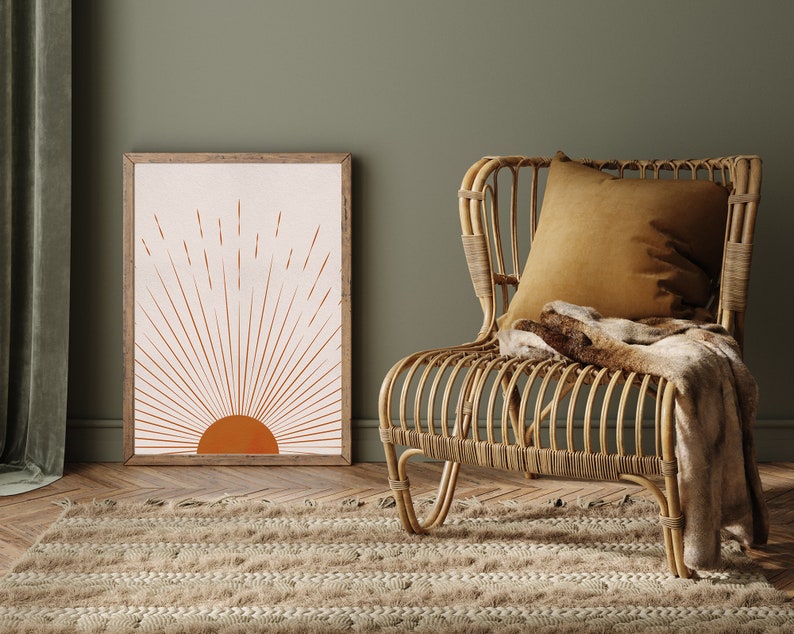 May include: A framed art print featuring a minimalist sunburst design in shades of orange and beige. The artwork is displayed next to a woven rattan chair with a mustard-colored pillow and a faux fur throw. A beige rug and green wall complete the scene.