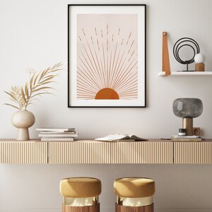 May include: A framed art print featuring a minimalist sunburst design in shades of orange and beige. The artwork is displayed above a wooden console with decorative elements, including a vase with dried foliage, books, and a modern lamp. Two gold and wood stools are in front of the console.