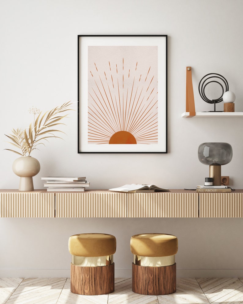 May include: A framed art print featuring a minimalist sunburst design in shades of orange and beige. The artwork is displayed above a wooden console with decorative elements, including a vase with dried foliage, books, and a modern lamp. Two gold and wood stools are in front of the console.