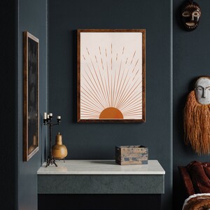 May include: A framed art print featuring a minimalist sunrise design in shades of orange and cream. The artwork is displayed on a dark teal wall, alongside other decorative items, including a candle holder and a wooden box. The overall aesthetic is warm and inviting.