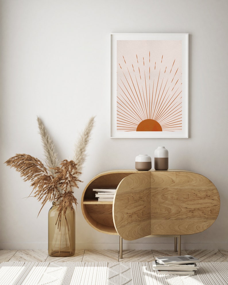 May include: A framed art print featuring a minimalist sunburst design in shades of orange and peach. The artwork hangs above a wooden cabinet with an oval shape and two small ceramic vases. A glass jar with dried pampas grass completes the decor.