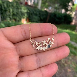 Puede incluir: Collar de oro con un dije de murciélago y el nombre "Danielle" escrito en cursiva.