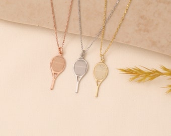 May include: Three tennis racket pendant necklaces in rose gold, silver, and gold. Each pendant features a detailed racket design, suspended from a delicate chain. The necklaces are displayed on a neutral-toned surface, with a sprig of wheat in the background.
