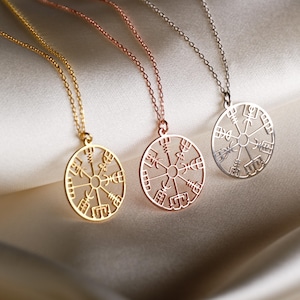 Puede incluir: Tres collares con un colgante circular que presenta un diseño de brújula con runas. Los collares son de oro, oro rosa y plata.