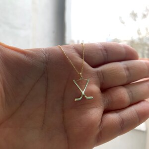 May include: A gold necklace with a pendant depicting crossed hockey sticks and a puck. The pendant hangs from a fine chain. The necklace is displayed against a blurred backdrop.