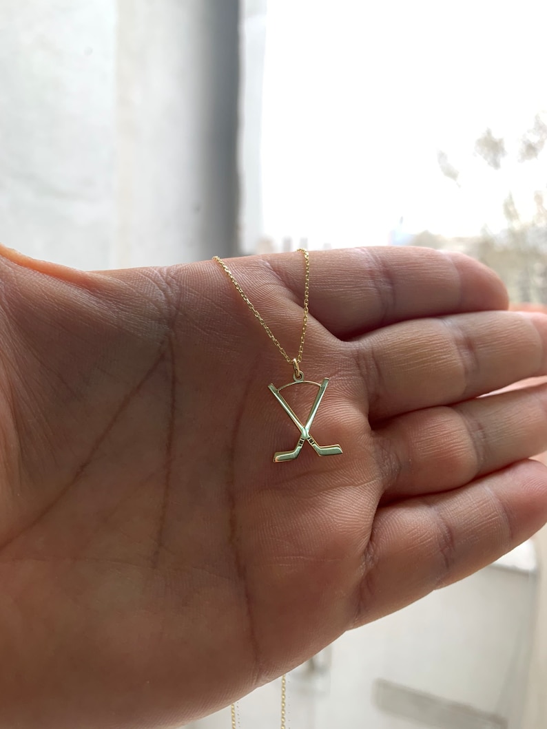 May include: A gold necklace with a pendant depicting crossed hockey sticks and a puck. The pendant hangs from a fine chain. The necklace is displayed against a blurred backdrop.