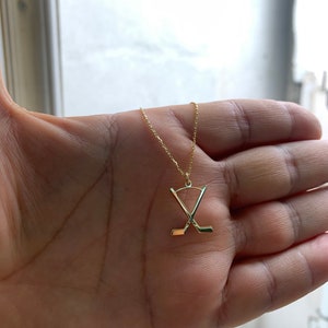 May include: A gold necklace with a hockey stick pendant. The pendant shows two crossed hockey sticks. The necklace is displayed on a hand, highlighting the fine chain and the pendant's design. The jewellery is likely for sports fans.