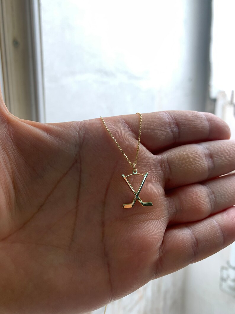 May include: A gold necklace with a hockey stick pendant. The pendant shows two crossed hockey sticks. The necklace is displayed on a hand, highlighting the fine chain and the pendant's design. The jewellery is likely for sports fans.