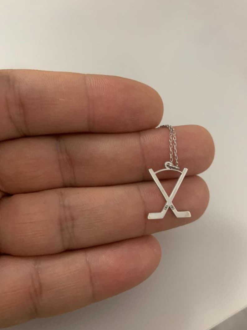 May include: A silver hockey stick pendant necklace. The pendant features two crossed hockey sticks with fine detailing. The necklace is on a delicate chain. The pendant is approximately 2 cm wide.