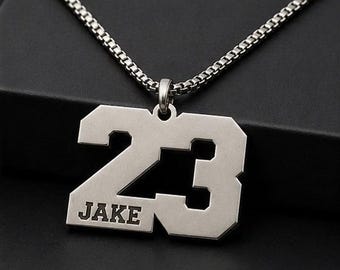 Puede incluir: Collar con colgante plateado con el número 23 y el nombre "JAKE". El colgante tiene forma rectangular con detalles recortados y cuelga de una cadena a juego. El collar se muestra sobre una superficie oscura.