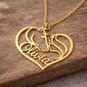 Puede incluir: Collar con colgante dorado con diseño en forma de corazón. El colgante presenta la silueta de una cabeza de caballo y el nombre "Olivia" en cursiva. La cadena delicada se apoya sobre un fondo de madera.