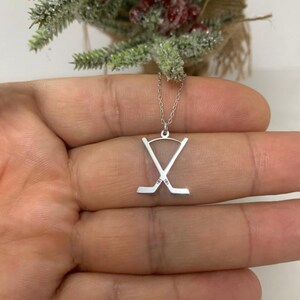 May include: A silver hockey stick pendant necklace. The pendant shows two crossed hockey sticks. The necklace is presented against a blurred background of a small decorative tree.