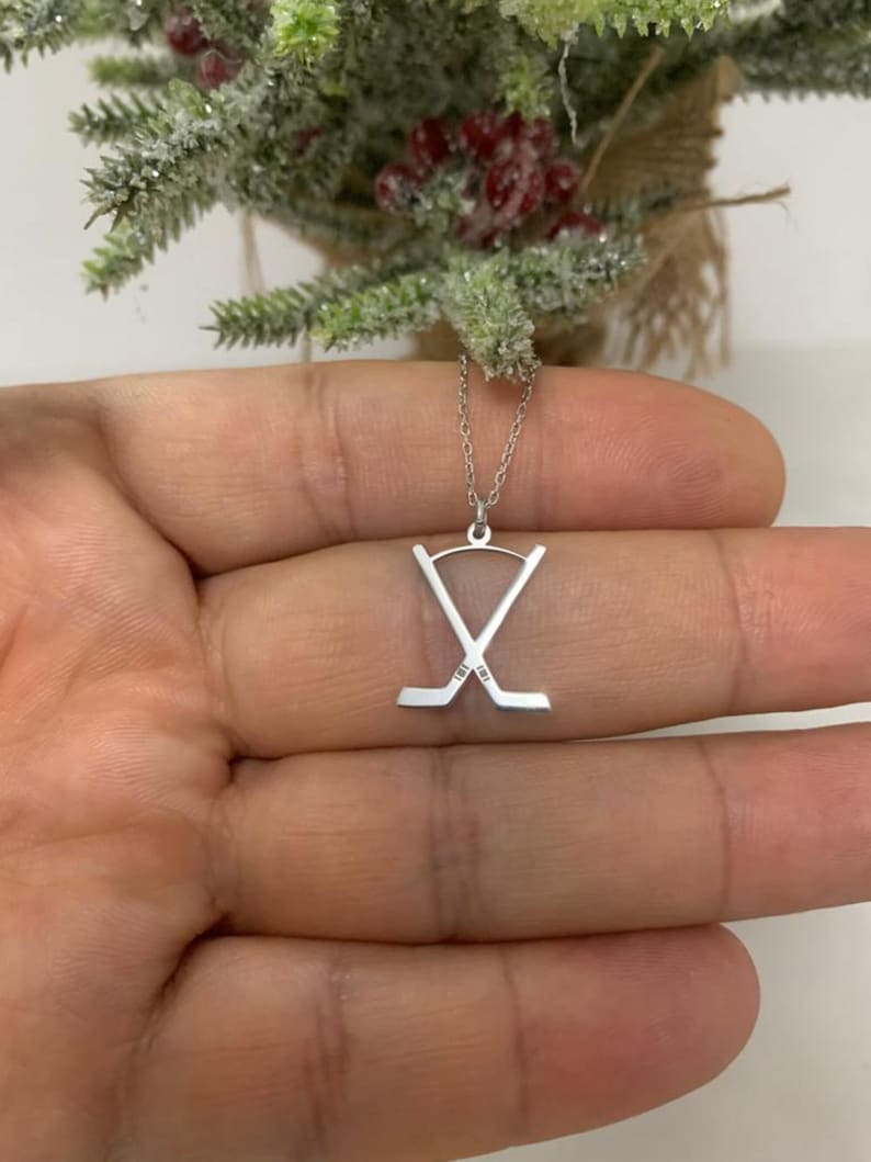 May include: A silver hockey stick pendant necklace. The pendant shows two crossed hockey sticks. The necklace is presented against a blurred background of a small decorative tree.