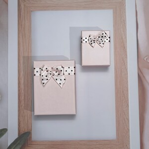 May include: Two small, rectangular, blush-coloured gift boxes with black polka dot bows and ribbons. The boxes are arranged on a light grey surface, inside a wooden picture frame. The bows are tied on top of the boxes.