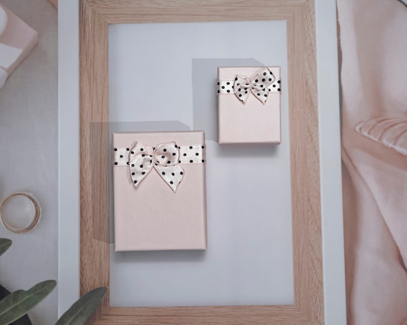 May include: Two small, rectangular, blush-coloured gift boxes with black polka dot bows and ribbons. The boxes are arranged on a light grey surface, inside a wooden picture frame. The bows are tied on top of the boxes.