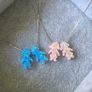 Puede incluir: Cuatro collares con colgantes en forma de persona. Dos colgantes son azules y dos son rosas. Los colgantes están hechos de un material iridiscente. Los collares tienen delicadas cadenas de plata.