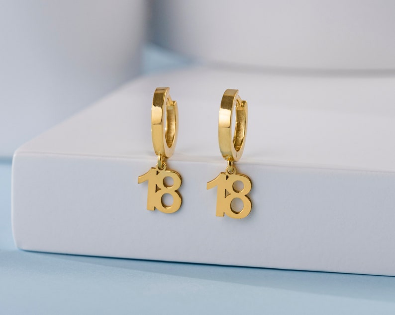 May include: A pair of gold hoop earrings with a dangling charm of the number 18.