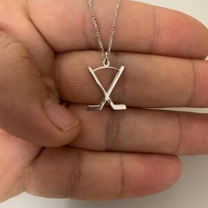May include: A silver hockey stick pendant necklace. The pendant shows two crossed hockey sticks. The necklace is presented against a plain background.