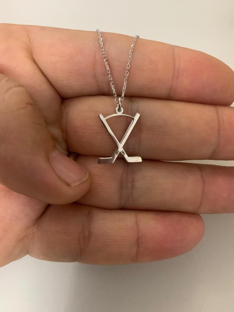May include: A silver hockey stick pendant necklace. The pendant shows two crossed hockey sticks. The necklace is presented against a plain background.
