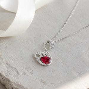 May include: A silver-toned necklace featuring a swan-shaped pendant. The swan is encrusted with small, clear, sparkling stones and features a prominent, pear-shaped, deep red gemstone set within the body of the swan design.