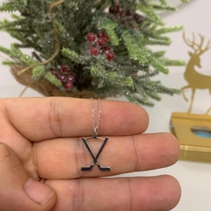 May include: Silver hockey stick pendant necklace. The pendant shows two crossed hockey sticks. The necklace is held in a hand, with a blurred background of a small Christmas tree and a gold reindeer decoration.
