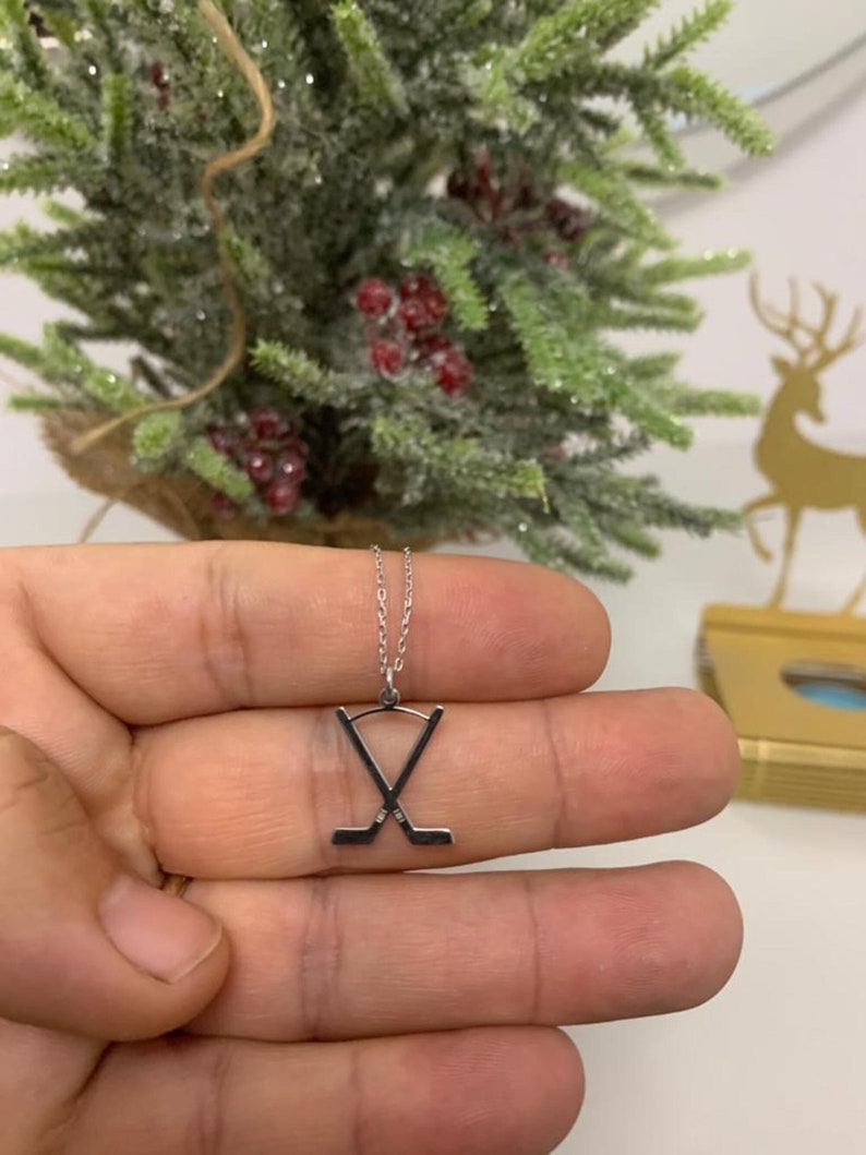 May include: Silver hockey stick pendant necklace. The pendant shows two crossed hockey sticks. The necklace is held in a hand, with a blurred background of a small Christmas tree and a gold reindeer decoration.