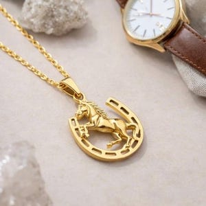以下が含まれることがあります： 金色のネックレス。馬のペンダントが蹄鉄の中に入っています。背景には、茶色の革バンドが付いた金色の腕時計があります。時計の文字盤は白です。ネックレスは明るい色の表面にあります。