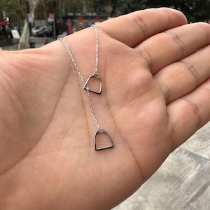 Puede incluir: Un collar de plata con dos pequeños colgantes en forma de estribo. El collar es delicado y fino.