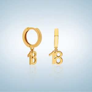 May include: A pair of gold hoop earrings with a dangling charm of the number 18.