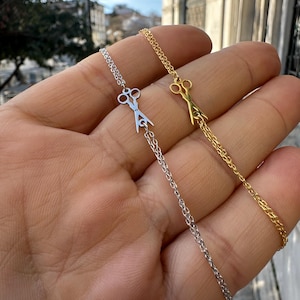 以下が含まれることがあります： 金と銀の繊細なチェーン2本に、それぞれハサミのペンダントがついたもの。チェーンは重ねてあり、ハサミは互いに向き合っている。