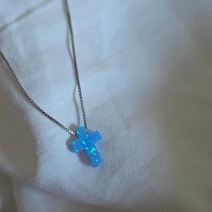 May include: A silver necklace with a blue cross pendant. The cross is a light blue color with hints of green. The necklace is displayed on a white, textured surface.