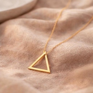 Puede incluir: Un collar dorado con un colgante triangular. El colgante es una forma geométrica simple suspendida de una delicada cadena. El collar se muestra sobre una tela beige suave, creando una estética minimalista.