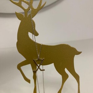 May include: A gold-coloured metal deer-shaped jewellery holder with a silver necklace. The necklace features a pendant with crossed hockey sticks. The deer has detailed antlers and stands on a rectangular base. The background is a plain white wall.