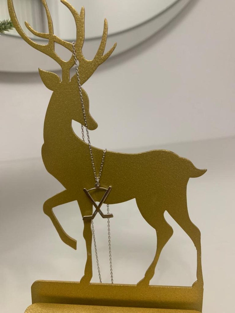 May include: A gold-coloured metal deer-shaped jewellery holder with a silver necklace. The necklace features a pendant with crossed hockey sticks. The deer has detailed antlers and stands on a rectangular base. The background is a plain white wall.